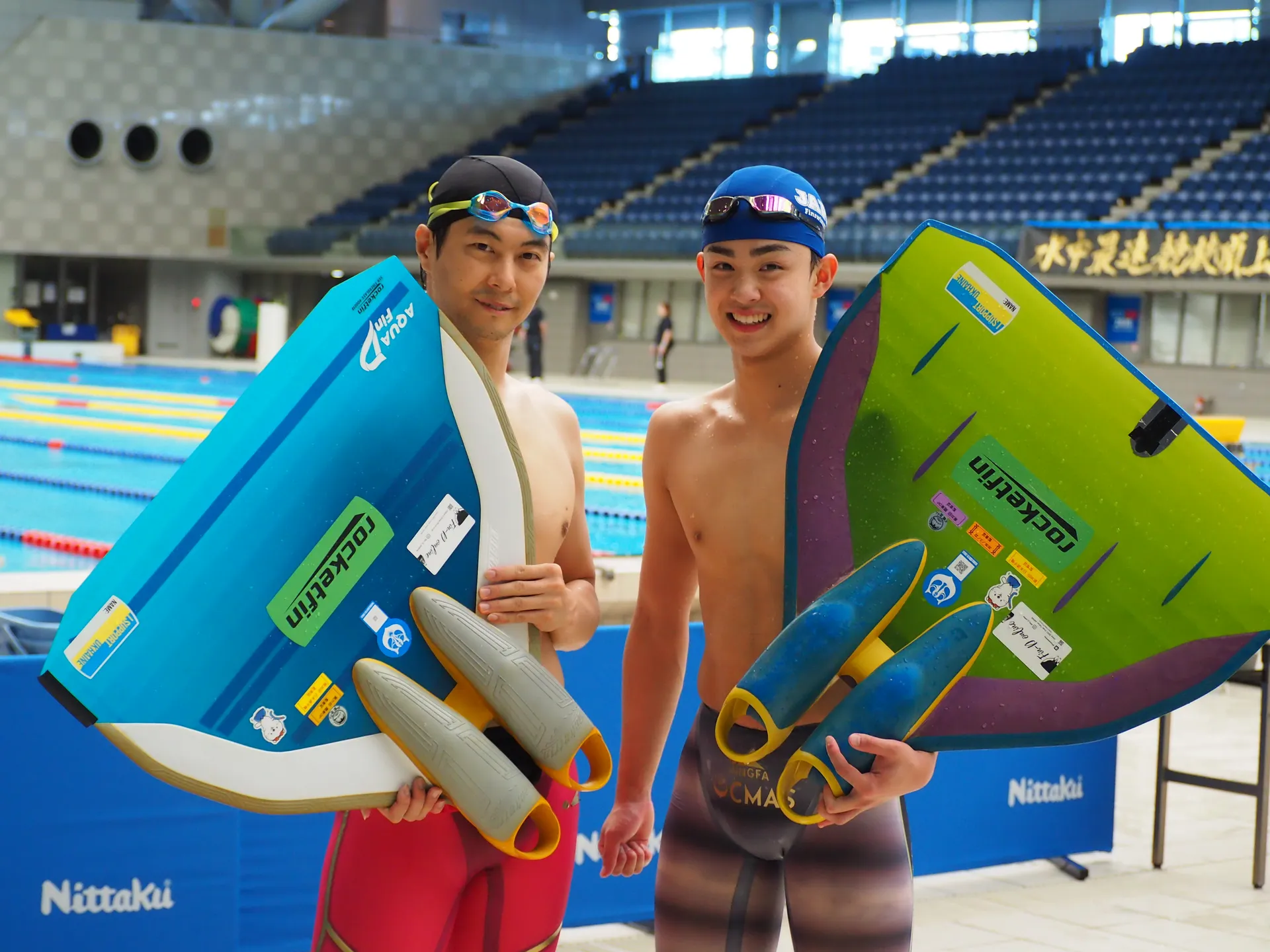 フィンスイミング選手2名がモノフィンとロケットフィンを持ちプールサイドで笑顔のポーズ