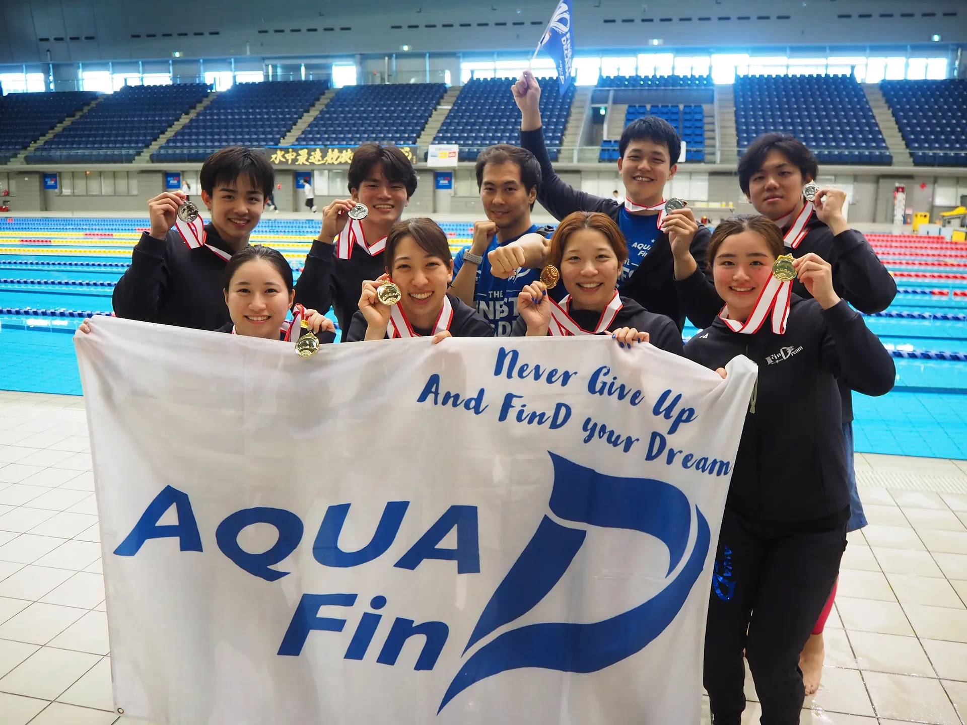 AQUA FinDのメンバーがメダルとチーム旗を掲げてプールサイドで記念撮影